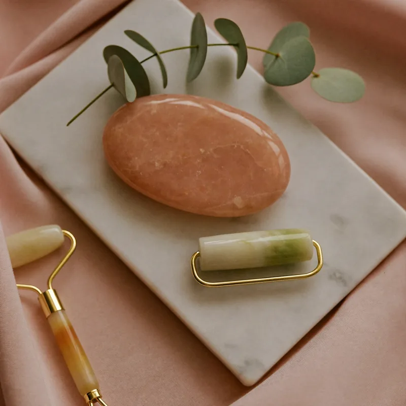 Rose quartz gua sha stone and jade facial roller on a marble tray with eucalyptus