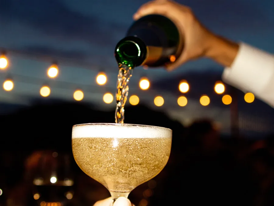 Champagne poured into a coupe glass with string lights glowing in the dusk behind