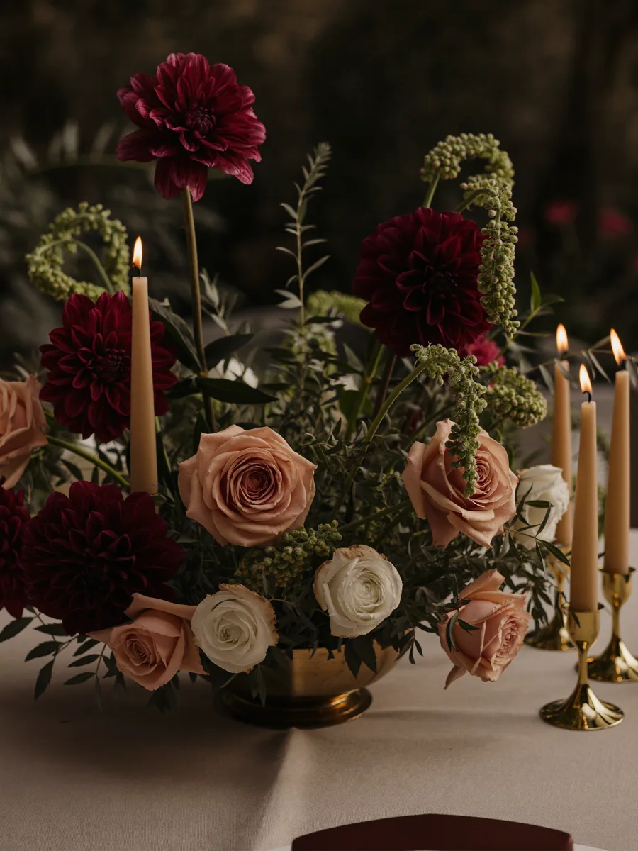 Lush centerpiece of burgundy dahlias, blush roses and taper candles in gold candlesticks